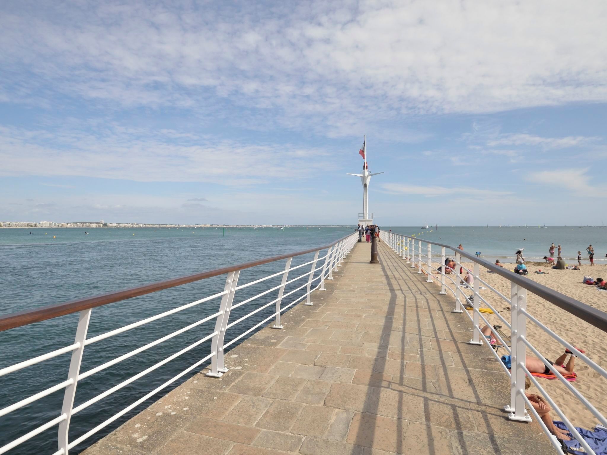 A pier with a white railing, a flagpole, and a beach with people.