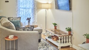 Una sala de estar con un sofá, una mesa de centro, una lámpara de pie, un televisor y plantas en macetas.