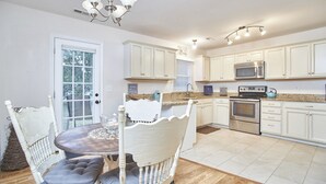 Una cocina con armarios blancos, encimera de granito, horno de acero inoxidable y microondas. Hay una zona de comedor con mesa de madera y sillas blancas.