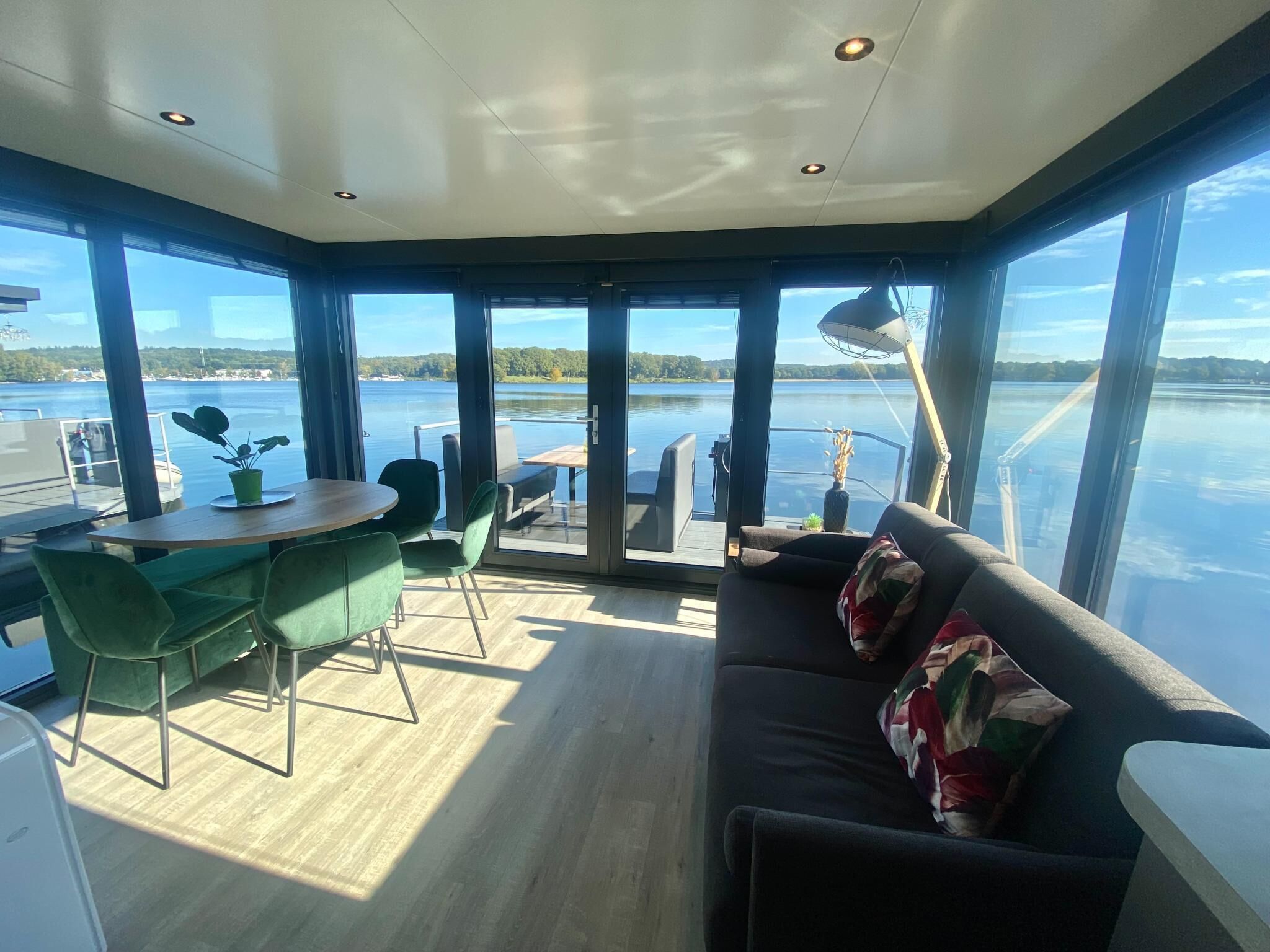 A modern interior with a dining area, a sofa, and large windows offering a view of a lake.