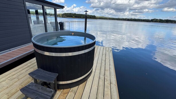 A hot tub on a wooden deck with a view of a calm lake and distant trees.