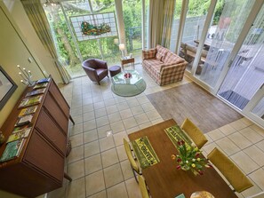 A spacious living area with a sofa, armchair, and coffee table, a wooden cabinet with magazines, and a view of greenery through large windows. - Wellness and tranquillity in green Twente (Oldenzaal)