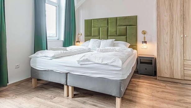 Un lit bien fait, avec une literie blanche, une tête de lit recouverte d’un tissu vert et une table de chevet avec une lampe.