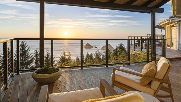 Une terrasse en bois offrant une vue sur la mer et le coucher de soleil. On y trouve deux chaises longues et un jardinier avec des plantes vertes.