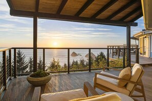 Une terrasse en bois offrant une vue sur la mer et le coucher de soleil. On y trouve deux chaises longues et un jardinier avec des plantes vertes.