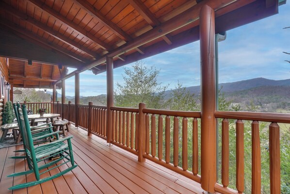 Una terraza de madera con barandilla, mesas y sillas, con vistas a un paisaje montañoso.
