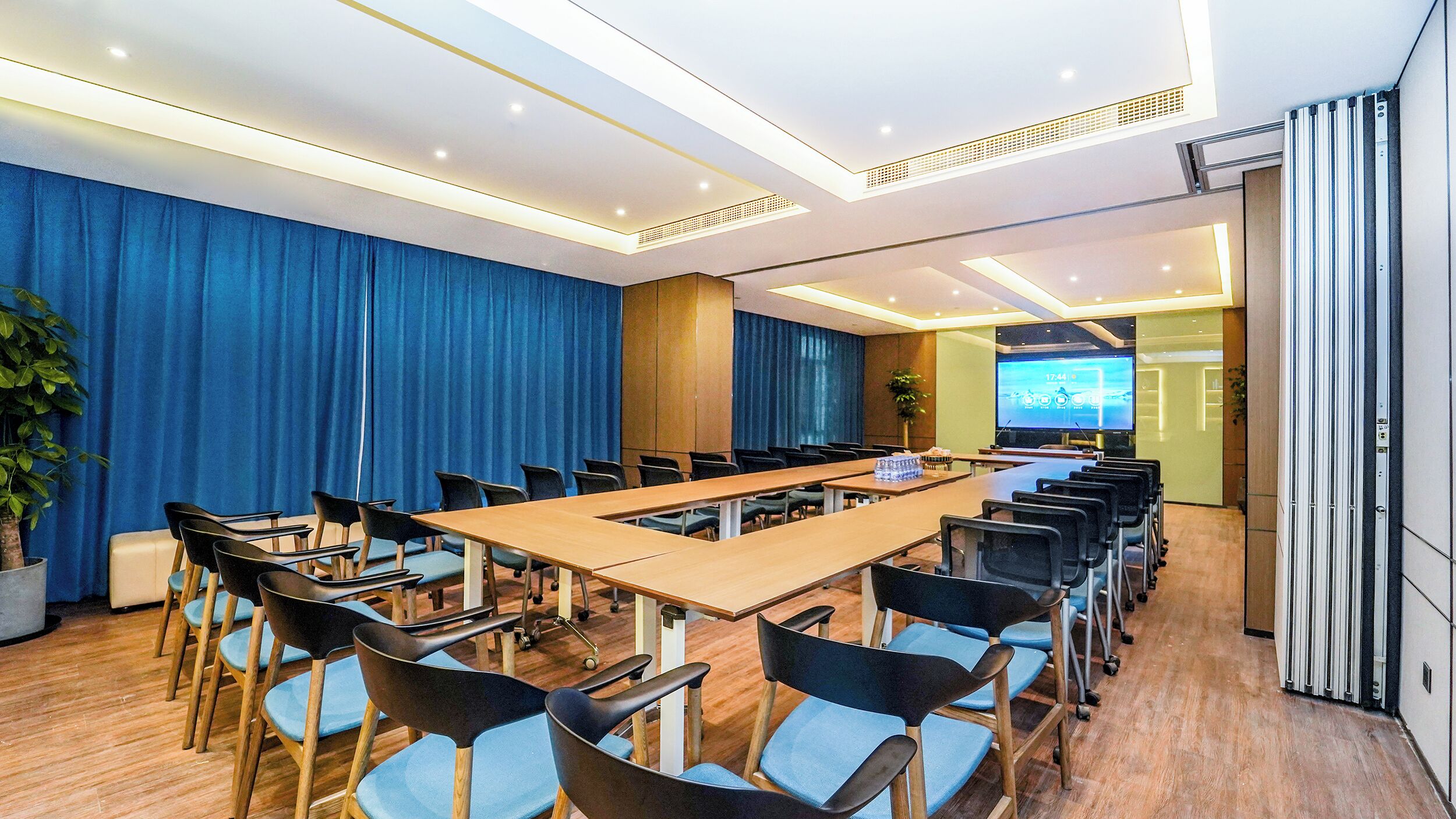 A conference room with a long table, chairs, a projector screen, and a blue curtain backdrop.