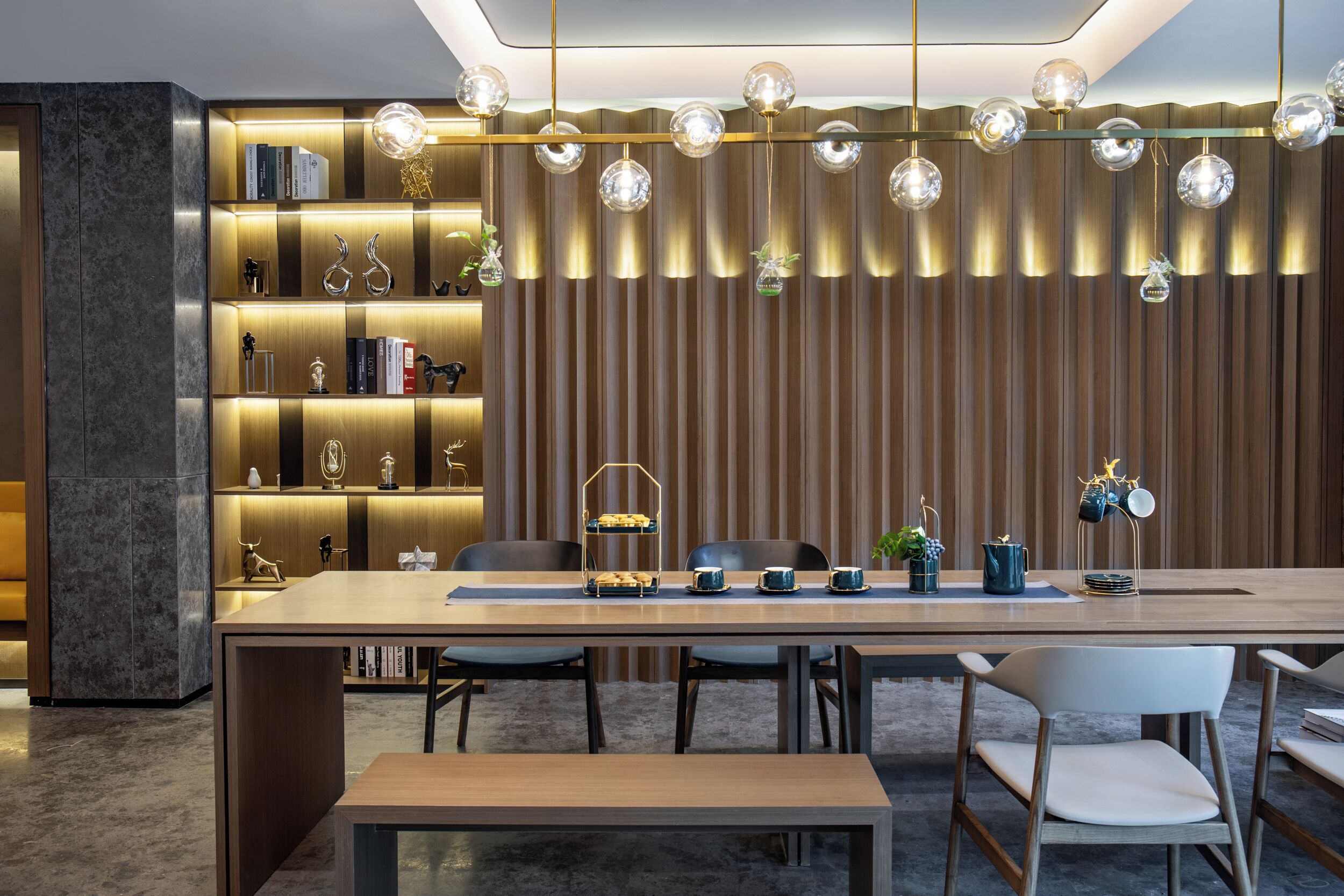 Un espace repas moderne avec une longue table, des chaises et une bibliothèque agrémentée d’objets décoratifs.