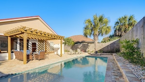 Un patio trasero con piscina, un área de patio techada con mobiliario y una cerca de madera.