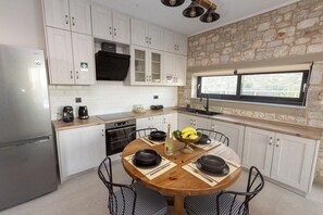 A modern kitchen with white cabinets, a stone accent wall, a dining table with black plates, and a window with a view. - Pierres Blanches Residences 2, with private pools (Mitilini)