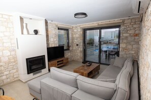 A modern living room with a stone wall, a flat-screen TV, a wooden coffee table, and a grey sofa. - Pierres Blanches Residences 2, with private pools (Mitilini)