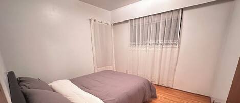 A bedroom with a bed, a window with a patterned curtain, and a wooden floor.