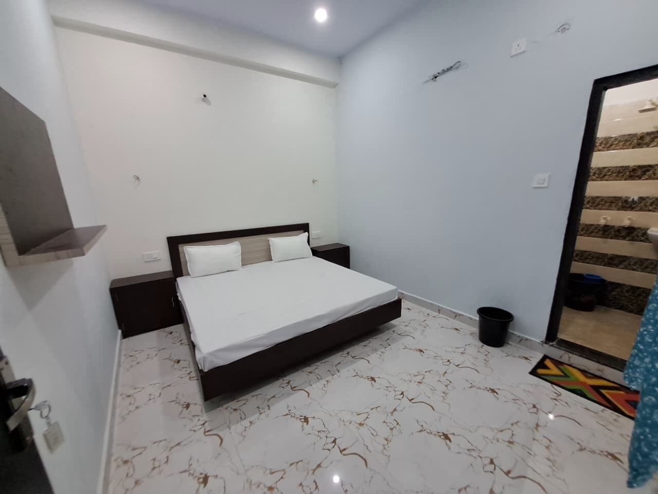 A bedroom with a bed, a nightstand, a wall-mounted shelf, a door, and a tiled floor.