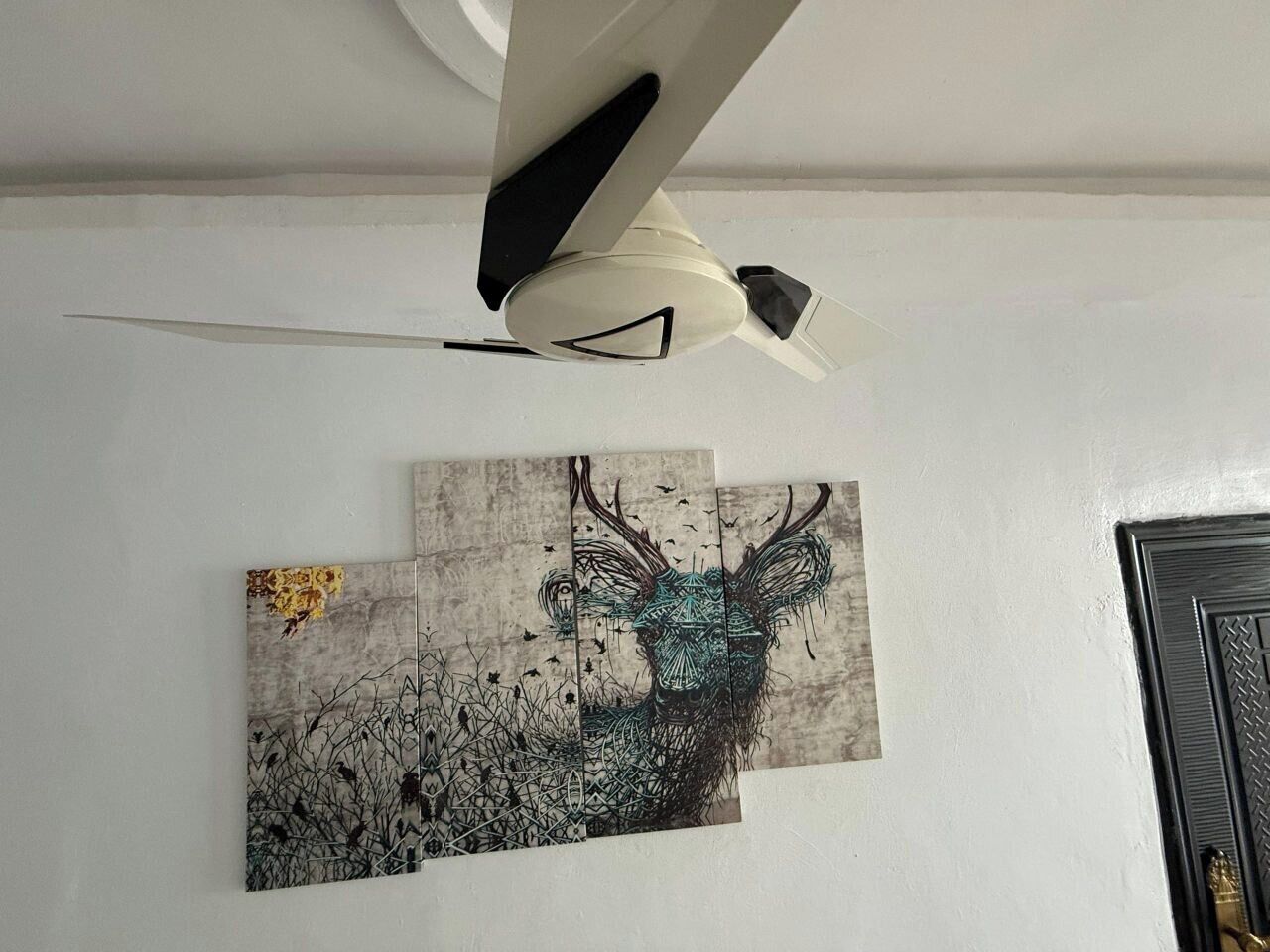 Een plafondventilator met een zwart-wit ontwerp, een aan de muur hangend kunstwerk van een hert, en een deur met een reliëfpatroon.