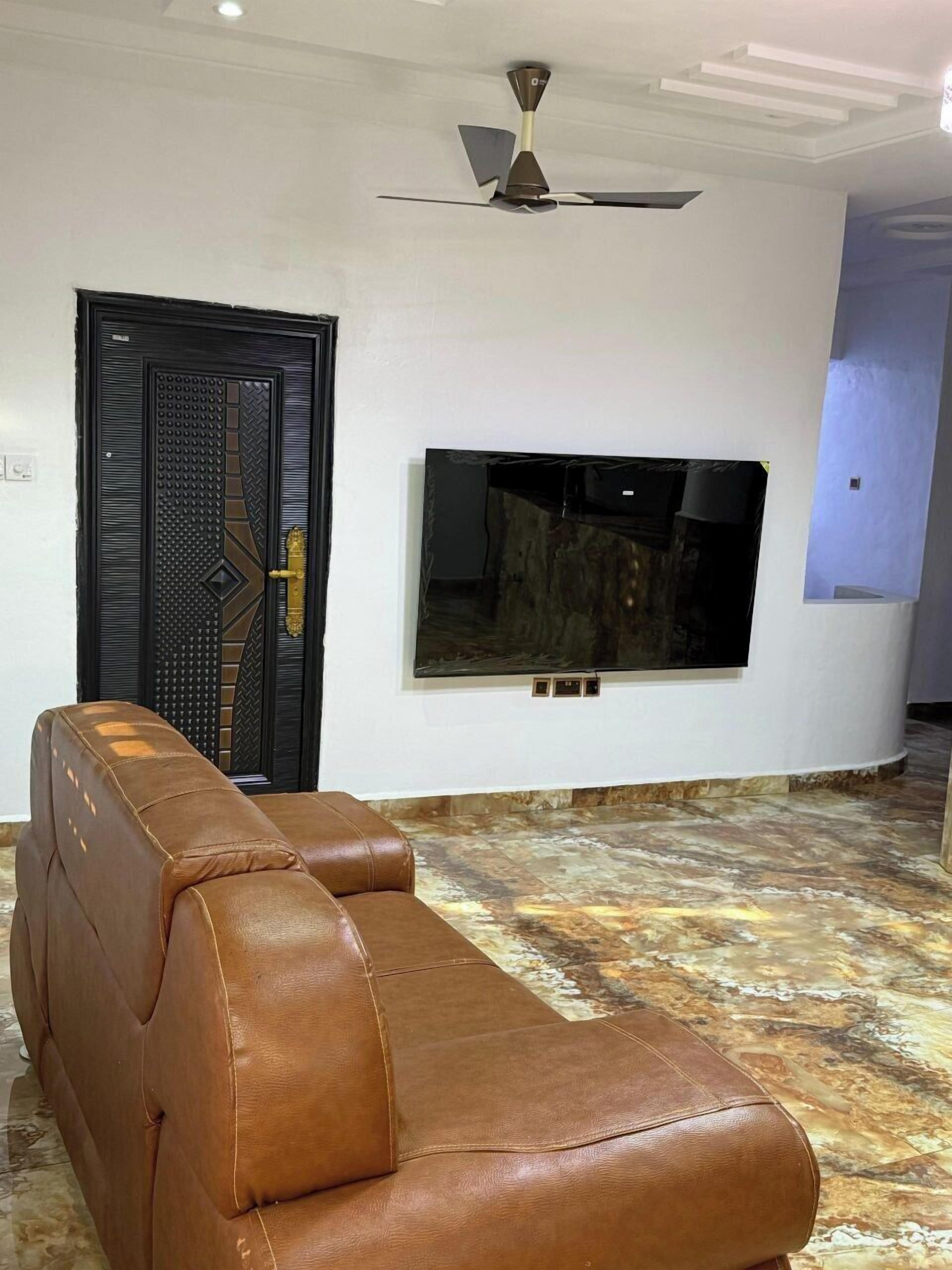 Ein Wohnzimmer mit einem braunen Ledersofa, einer schwarzen Tür mit dekorativem Muster, einem Deckenventilator und einem flachbildfernseher an der Wand.