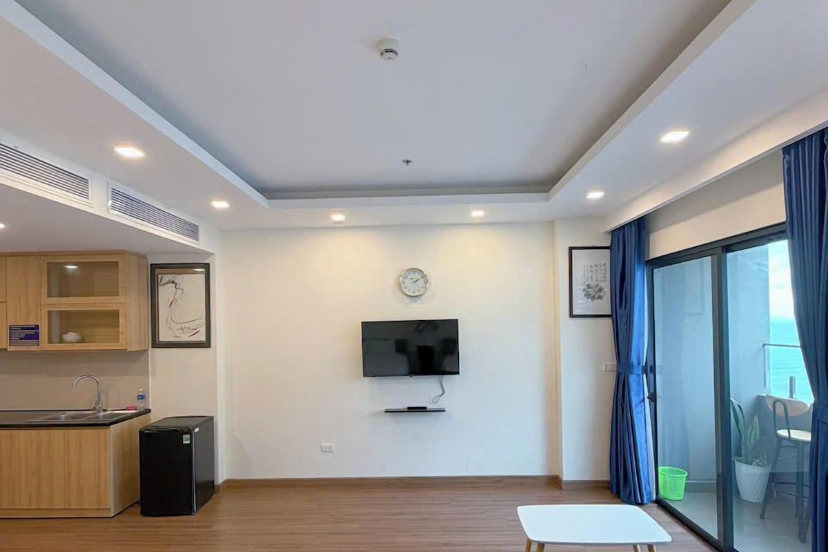 A modern hotel room with a kitchenette, a flat-screen TV, a sofa, and a large window with curtains.