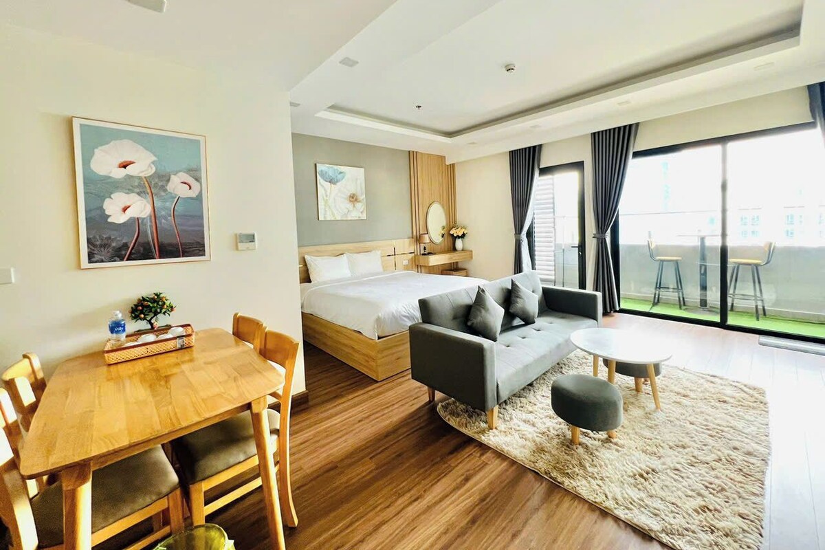 A modern hotel room with a wooden dining table, a sofa, a round coffee table, and a bed with a wooden headboard.