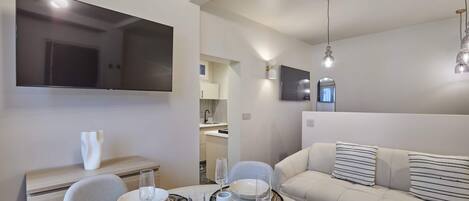 A modern living room with a sofa, dining table set with plates and glasses, and a flat-screen TV mounted on the wall.
