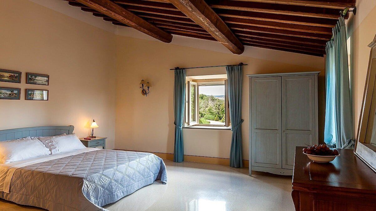 Quarto com cama, mesa de cabeceira, janela com cortinas, guarda-roupa e vista para o verde lá fora.