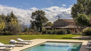 Piscina com espreguiçadeiras, um edifício de pedra e uma área gramada com árvores.