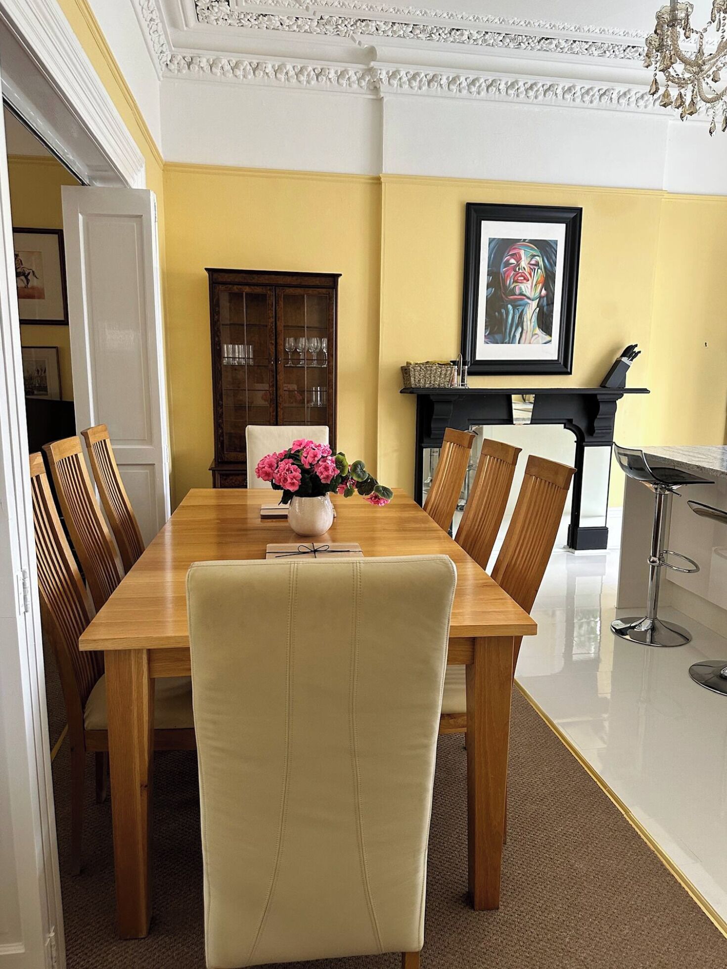 Uma sala de jantar com mesa de madeira, cadeiras, um vaso de flores, um armário e um quadro na parede.