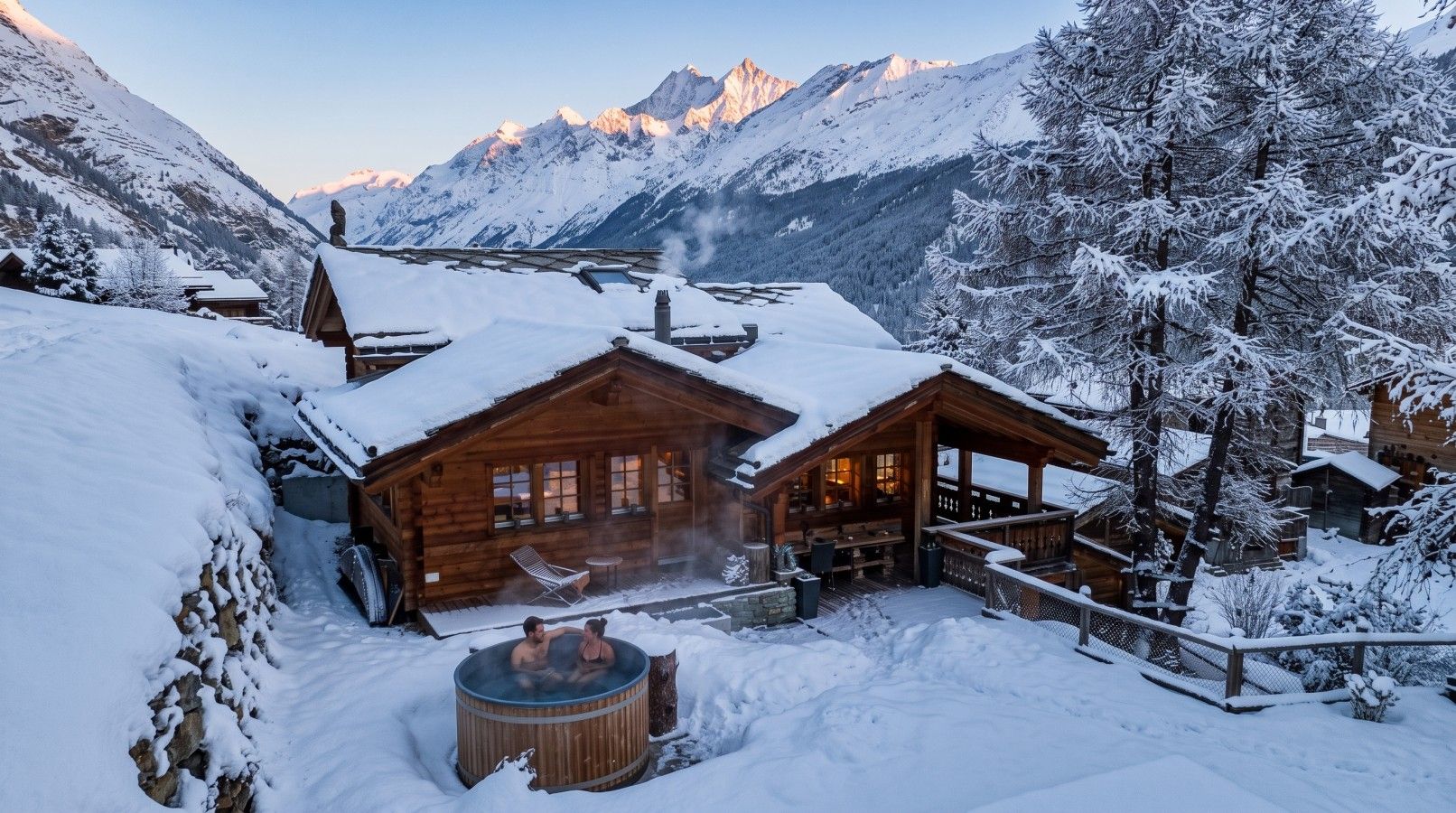 A wooden cabin with a hot tub in a snowy landscape, surrounded by mountains.