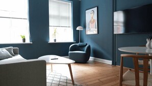 A modern living room with a blue wall, a round white table, a grey sofa, a blue armchair, and a floor lamp. - Greenbrook Blue Suites (Aalborg)