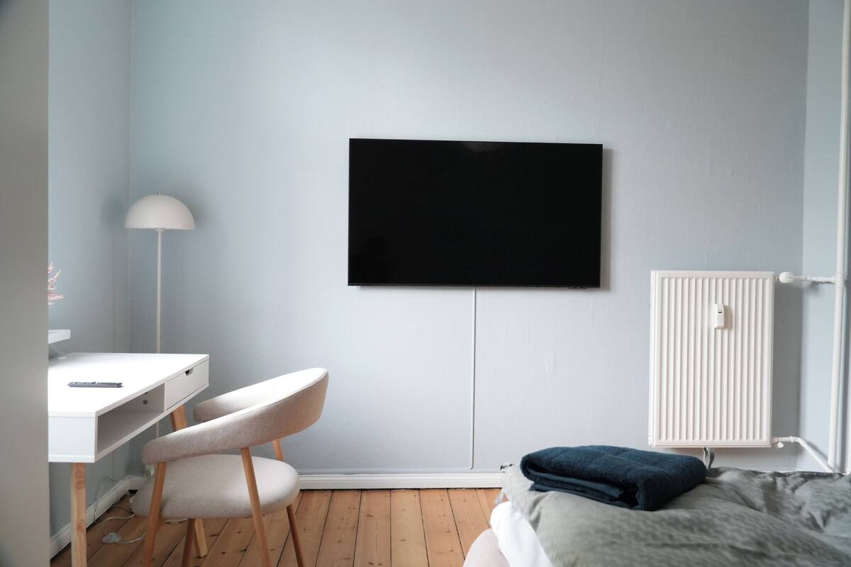 A bedroom with a bed, a desk with a chair, a wall-mounted TV, and a radiator.