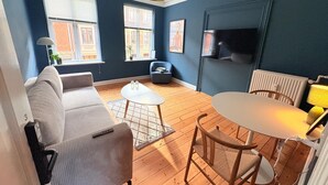 A modern living room with a sofa, a coffee table, a dining table with chairs, a TV, and large windows with blinds. - Greenbrook Blue Suites (Aalborg)