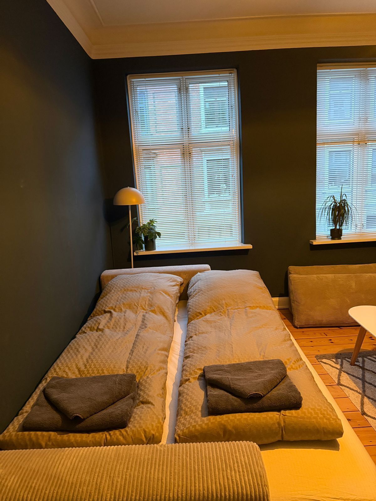 Una habitación con cama, dos ventanas con persianas, una mesita y una lámpara.