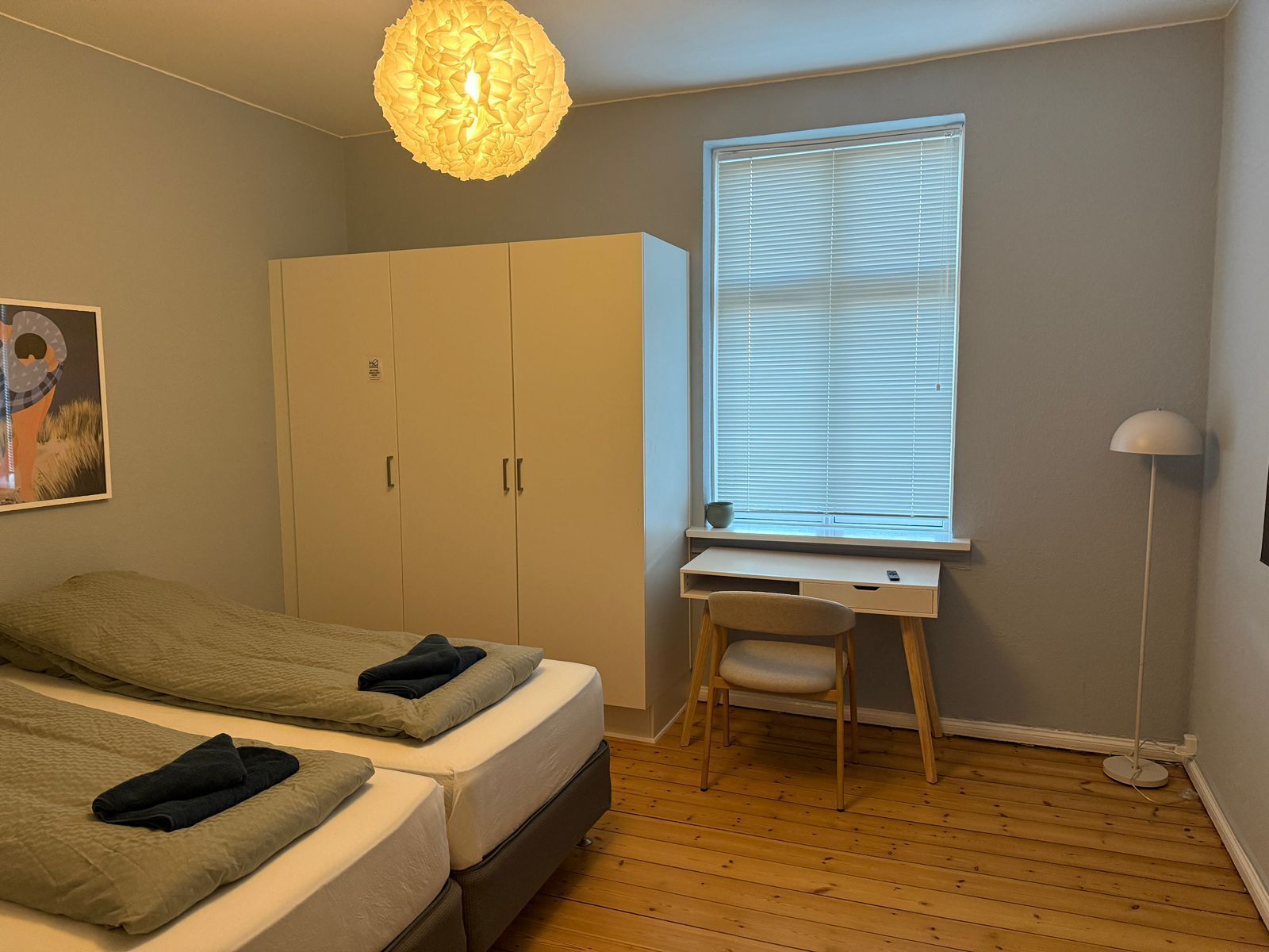 A bedroom with two beds, a desk, a chair, a wardrobe, and a window with blinds.