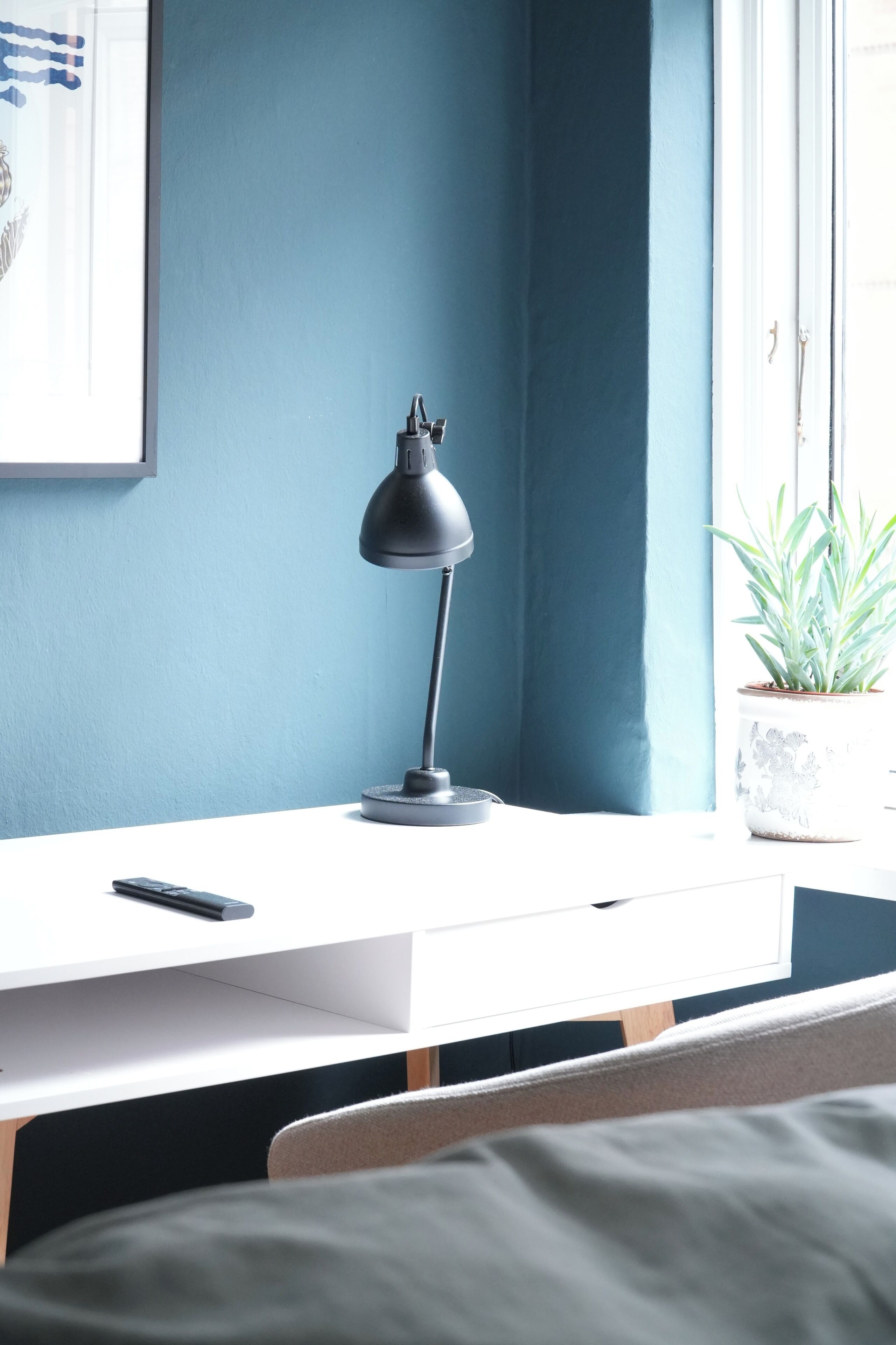 A desk with a lamp, a potted plant, and a framed picture on the wall.