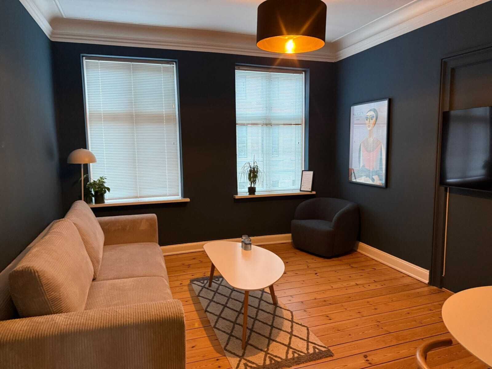 Una sala de estar con sofá, una mesita, una silla y televisor. Hay dos ventanas con persianas y un candelabro colgante.
