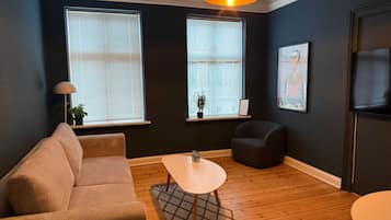 Una sala de estar con sofá, una mesita, una silla y televisor. Hay dos ventanas con persianas y un candelabro colgante.