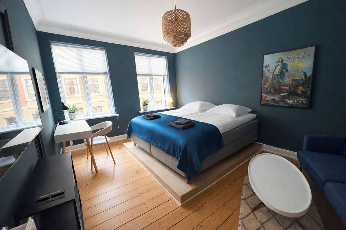 A bedroom with a bed, a desk, a chair, a round table, and a painting on the wall.