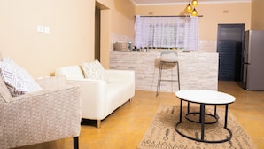 A living room with a white sofa, a patterned armchair, a small round table, and a kitchen area with a refrigerator and a window with curtains. - Pabvute 8859 Apartments (Victoria Falls)