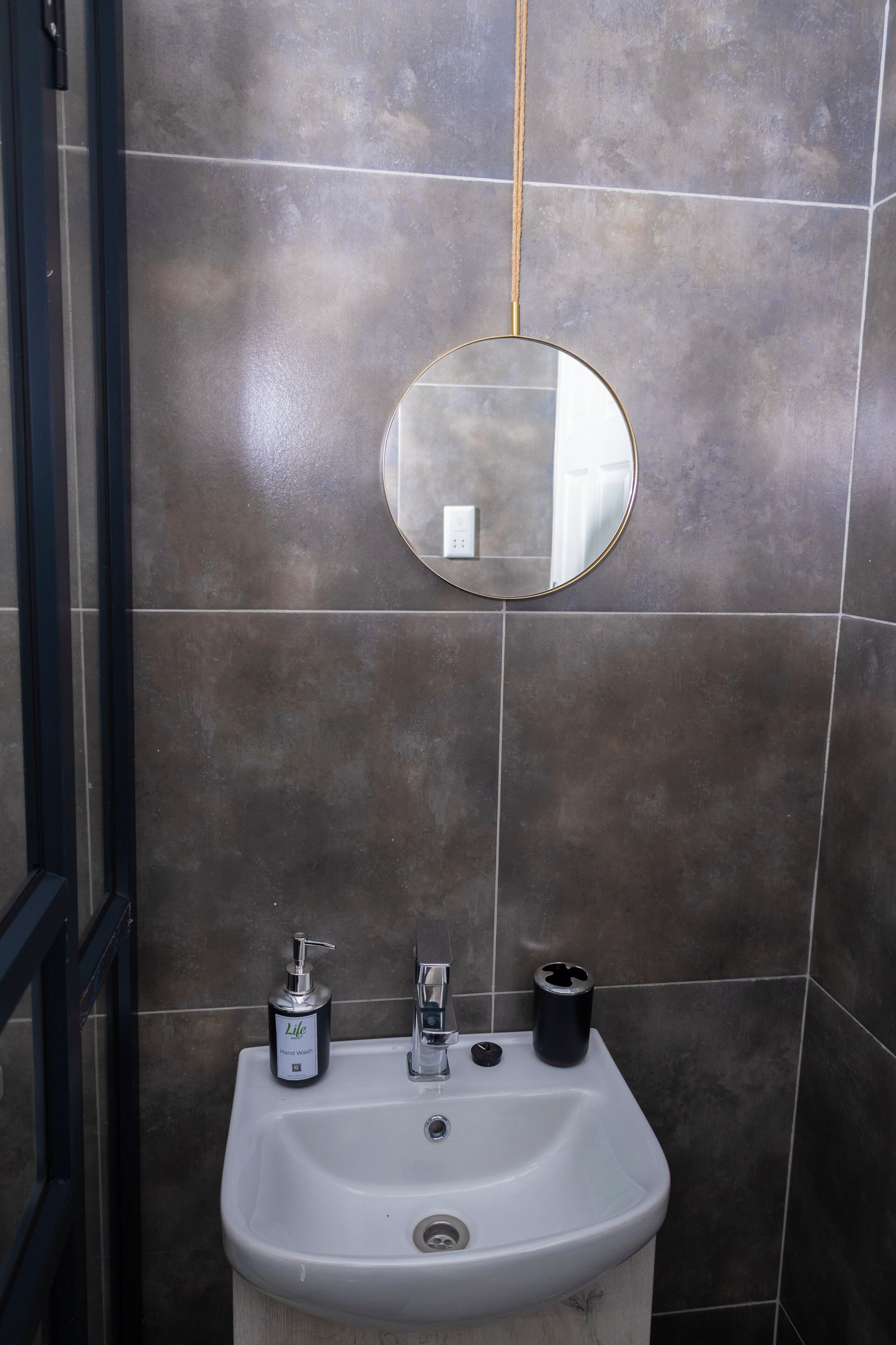 A bathroom with a white sink, a round mirror, a bottle of hand soap, and a black trash can.