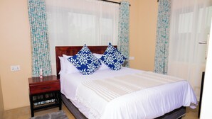 A neatly made bed with white bedding and blue floral pillows, a wooden nightstand with a drawer, and a grey rug on the floor. - Pabvute 8859 Apartments (Victoria Falls)