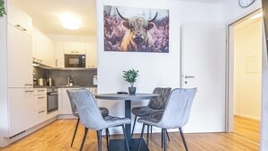 Ett modernt kök med vita skåp, en mikrovågsugn och en kaffemaskin. Det finns en matplats med ett runt bord och två grå stolar. En krukväxt står på bordet, och en tavla med en tjur hänger på väggen.
