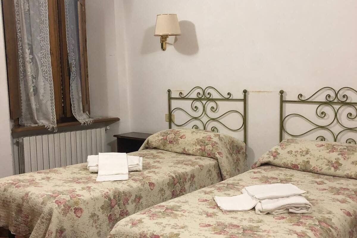 A room with two single beds, each with floral bedding and a metal headboard. There is a wall-mounted lamp and a window with patterned curtains.