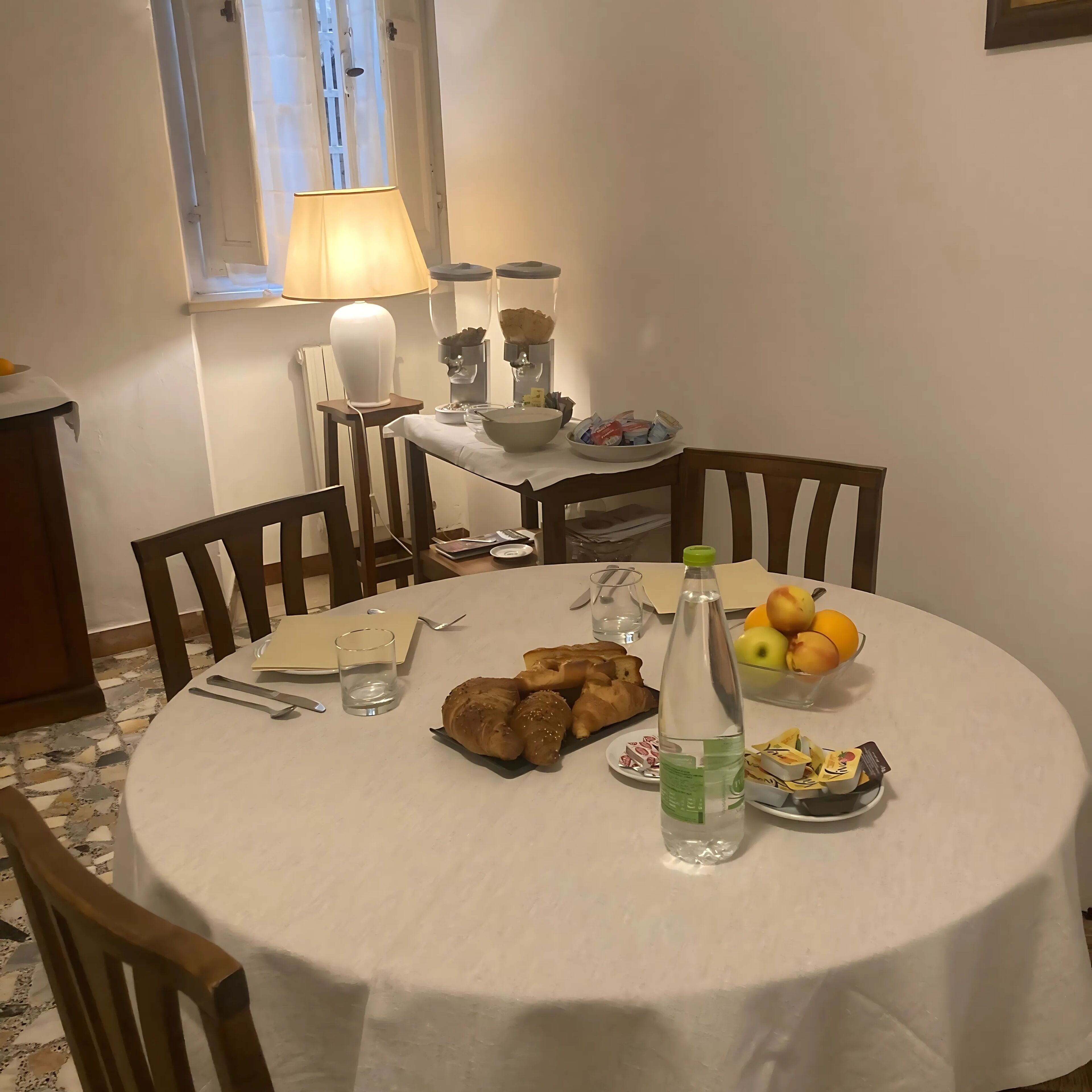 Una mesa redonda con mantel blanco, rodeada de sillas de madera. Sobre la mesa, hay croissants, una botella de agua, un cuenco de fruta y un plato con pasteles. Una lámpara está en la pared detrás de la mesa, y se ve una ventana con cortinas.