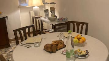 Une table de salle à manger ronde, recouverte d’une nappe blanche, est entourée de chaises en bois. Sur la table, on trouve des croissants, une bouteille d’eau, un saladier de fruits et une assiette de pâtisseries. Une lampe est fixée au mur derrière la table, et une fenêtre avec des rideaux est visible.