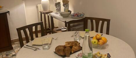 Una mesa redonda con mantel blanco, rodeada de sillas de madera. Sobre la mesa, hay croissants, una botella de agua, un cuenco de fruta y un plato con pasteles. Una lámpara está en la pared detrás de la mesa, y se ve una ventana con cortinas.