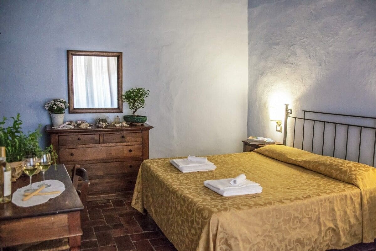 A hotel room with a bed, a wooden dresser, a mirror, a small potted plant, and a table with wine glasses and a placemat.