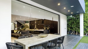 Un'area pranzo moderna con tavolo in legno e sedie nere, una scala che conduce a un piano superiore e una cucina visibile attraverso porte in vetro.