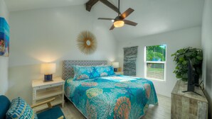 Une chambre à coucher comprenant un lit, une table de chevet, une chaise, un ventilateur de plafond, une fenêtre avec des stores et un élément de décoration fixé au mur.