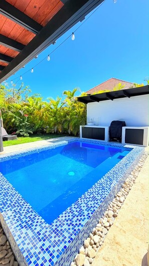 Ein Swimmingpool mit blauen Mosaikfliesen, umgeben von einem Kiesstreifen, unter einem überdachten Bereich mit Lichterketten.