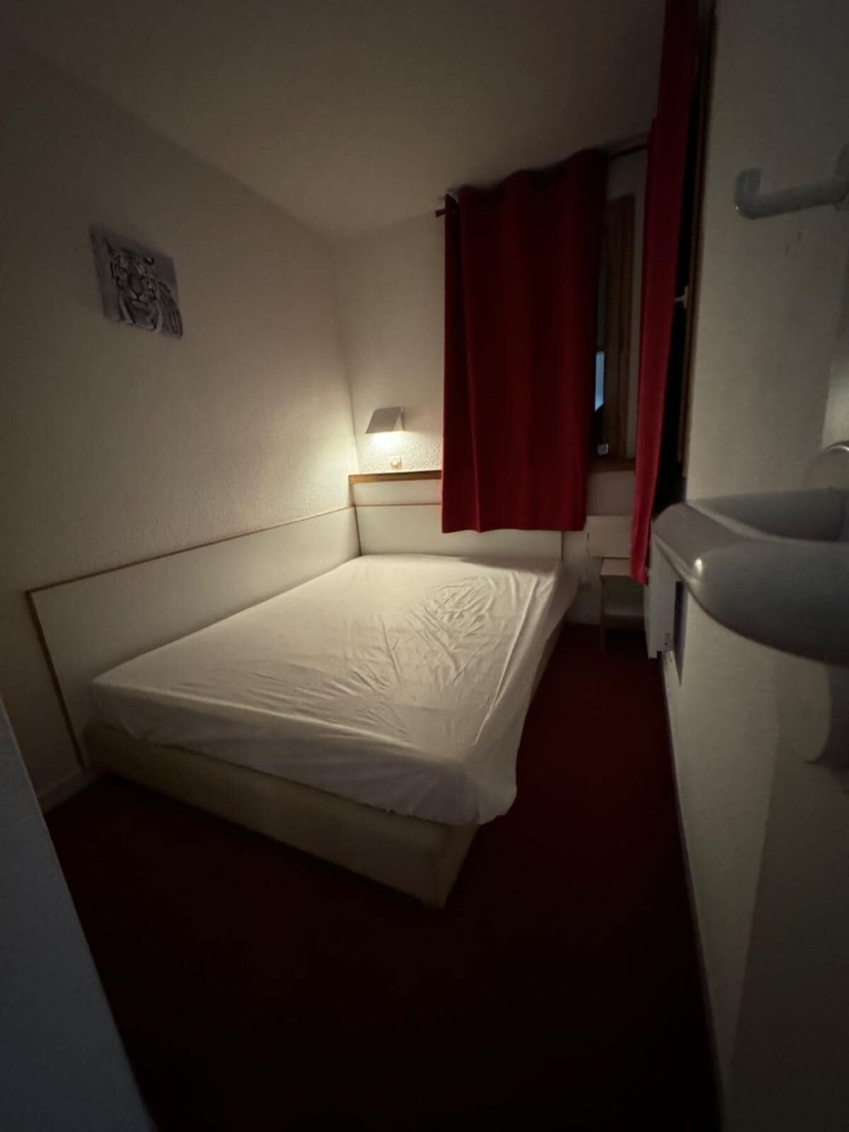 A small, dimly lit room with a single bed, a nightstand with a lamp, and a red curtain.