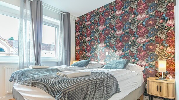 Une chambre avec un grand lit, un mur orné d’un motif floral, une fenêtre avec des rideaux et une table de chevet avec une lampe.