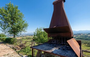 Ein rustikaler Grill im Freien mit Schornstein, umgeben von einem Holzzaun und einem Baum, mit Blick auf eine hügelige Landschaft.